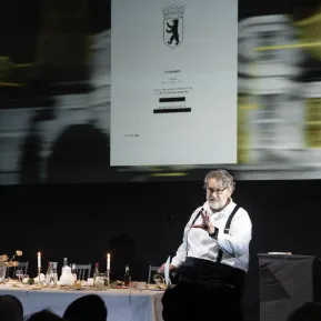 Andreas Beck liest aus geheimen Dokumenten, neben ihm eine lange, gedeckte Tafel