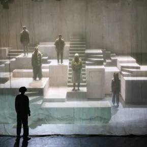 Ein Kind steht vor einem Vorhang. Dahinter sieht man schemenhaft ein Labyrinth mit weiteren Kindern