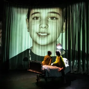 Zwei Menschen sitzen auf einem Krankenbett. Hinter ihnen eine Riesen große Projektion von einem Kind