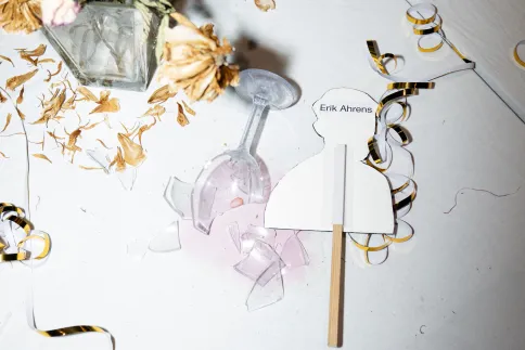 Ein zerbrochenes Glas liegt auf einem Tisch, daneben ein Schild mit der Aufschrift "Erik Ahrens"
