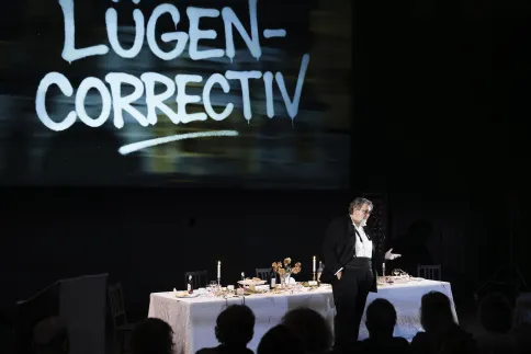 Andreas Beck steht vor einer gedeckten Tafel, hinter ihm ist "Lügen Correctiv" auf einer Leinwand projiziert