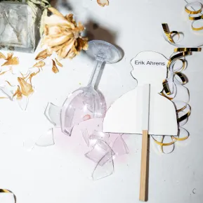 Ein zerbrochenes Glas liegt auf einem Tisch, daneben ein Schild mit der Aufschrift "Erik Ahrens"