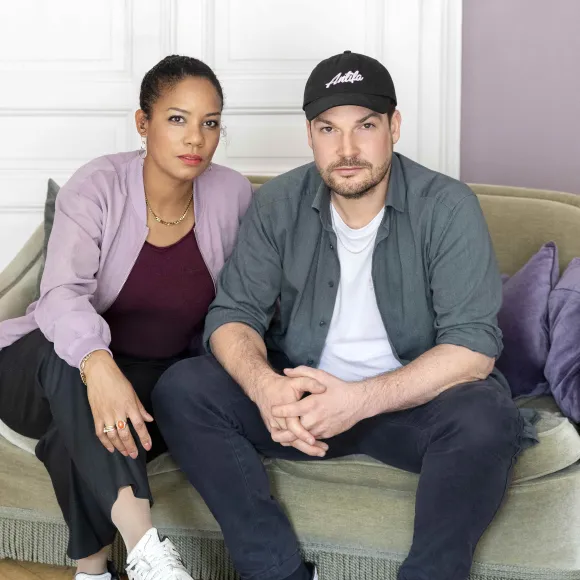 Hadija Haruna-Oelker und Max Czollek sitzen auf einem Sofa und schauen in die Kamera.